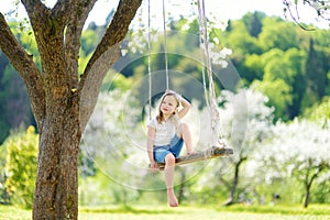 Cute little girl having fun on a swing in blossoming old apple tree garden outdoors on sunny spring day