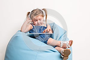 Cute little girl in headphones listening to music using a tablet and smiling while sitting on blue big bag