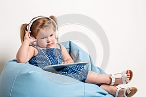 Cute little girl in headphones listening to music using a tablet and smiling while sitting on blue big bag