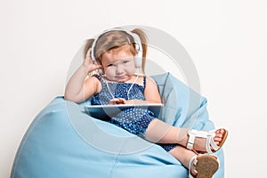 Cute little girl in headphones listening to music using a tablet and smiling while sitting on blue big bag