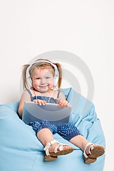 Cute little girl in headphones listening to music using a tablet and smiling while sitting on blue big bag