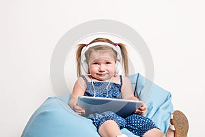 Cute little girl in headphones listening to music using a tablet and smiling while sitting on blue big bag