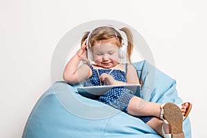 Cute little girl in headphones listening to music using a tablet and smiling while sitting on blue big bag