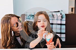 Cute little girl and her attractive cheerful mom wearing trendy sunglasses having fun at home and eating dessert