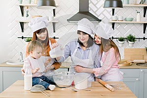 Cute Little Girl and Her Beautiful Mom, Aunt and Grandmother in Aprons and hats having fun While pouring milk to flour