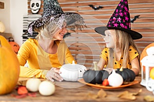 Cute little girl and her mother, both in witch costumes, sitting behind a table in Halloween theme decorated room.