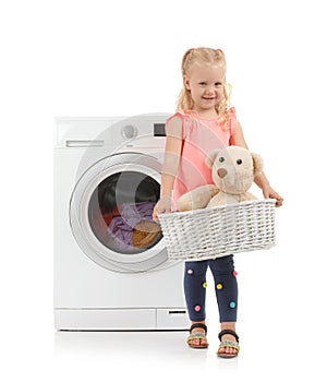 Cute little girl holding laundry basket with teddy bear near washing machine