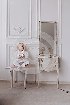 A cute little girl is holding a makeup brush and having fun at home. baby Girl is sitting on the chair near the classic mirror