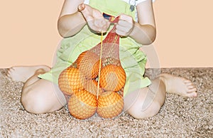 A cute little girl holding a net with big yellow oranges is sitting on the floor in the room. The child holds juicy, delicious, sw