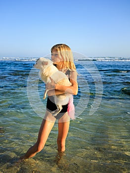 Cute little girl holding small white chihuahua dog with pink tail on the beach. Childhood concept. Spending time outdoor. Ocean
