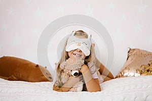 Cute little girl hugging a teddy bear is lying on a cozy bed, with a sleeping mask and smiling