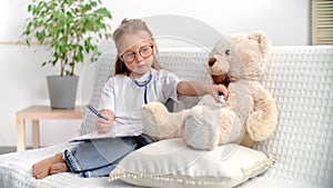 Cute little girl imagining playing doctor with bear toy use stethoscope writing prescription at home