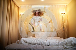 Cute little girl jumping on white bed in evening in sleeping room or in hotel