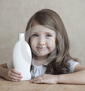 Cute little girl keeping white plastic shampoo bottle in her hands, looking at the camera. Baby bath, hygiene accessories