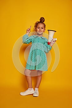 Cute little girl, kid wearing color dress shouting at megaphone isolated on yellow studio background. Concept of