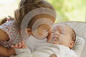 Cute little girl kissing his newborn sister. Toddler kid meeting new born sibling. Infant sleeping in white bouncer