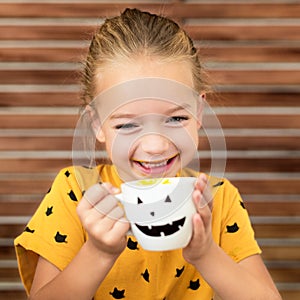 Cute little girl laughing and drinking pumpkin soup out of a cup with Halloween anthropomorphic smiley face. Halloween background.