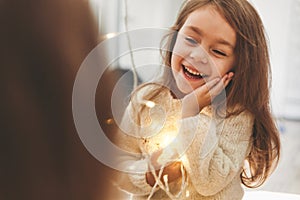 Cute little girl laughing and looking at morror with bright christmas garland in her hands