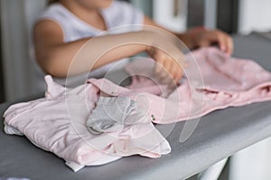 Cute little girl is leaning on ironing clothes on board at home. Daughter helping to mother ironing clothes for baby