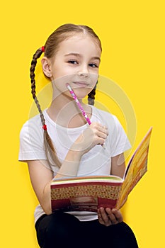 A cute little girl is learning lessons while holding a notebook and a pencil in her hand, or drawing while sitting on a yellow