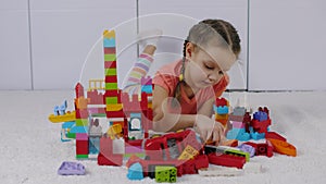 Cute little girl lies on carpet and playing with colorful lego building blocks