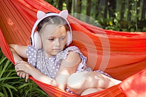 Cute little girl lies in a hammock and listening to music wearing wireless headphones at sunny summer day.