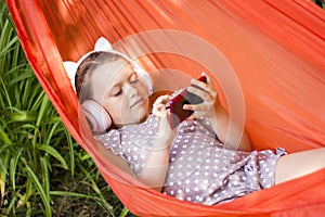 Cute little girl lies in a hammock with smartphone and wireless headphones and listening to music.