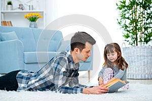 Cute little girl listening to dad reading fairy tale lying on warm floor together, caring father holding book , family hobbies