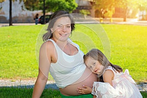 Cute little girl listening to her mothers pregnant belly in summer nature