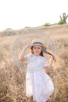 A cute little girl with long blond curly hair in a white summer dress and a straw boater hat in a field in the countryside