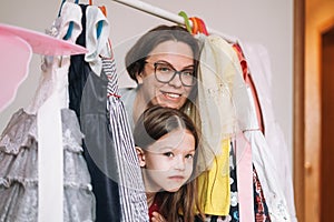 Cute little girl with long dark hair in red dress with mother young woman in glasses among her beautiful dresses in wardrobe in