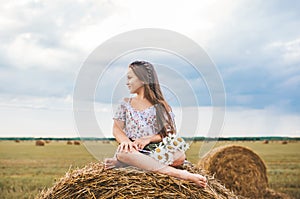 Cute little girl with long hair and with bouquet of chamomiles is sitting on a haystack in the field and enjoys sunset