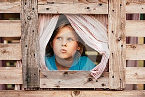 Cute little girl looks out window of wooden children's house. Holidays in village. Happy childhood.