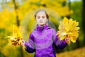 Cute little girl making funny faces on beautiful autumn day at city park. Funny child picking autumn leaves on sunny day.