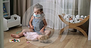 Cute little girl in medical mask with stethoscope playing doctor with toys at home floor.