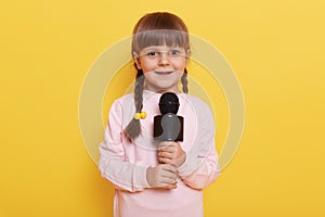 Cute little girl with microphone posing isolated on yellow color background, singing or telling poem, looks at camera with