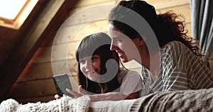 Cute little girl and mother lying on bed with smartphone