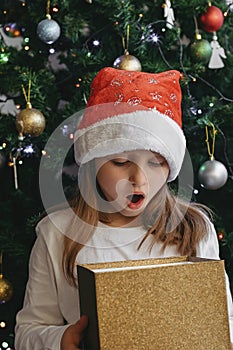 A cute little girl opens a Christmas present with surprise in a New Year`s hat under the tree. Holiday, present, miracle, magic