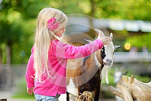 Cute little girl petting and feeding a goat at petting zoo. Child playing with a farm animal on sunny summer day.