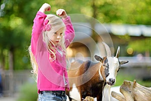 Cute little girl petting and feeding a goat at petting zoo. Child playing with a farm animal on sunny summer day.