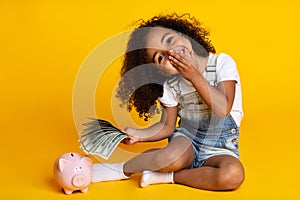 Cute little girl with piggy bank and banknotes, yellow background