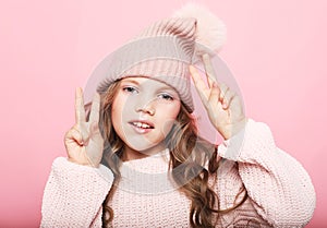 Cute little girl in pink sweater and winter hat showing victory sign
