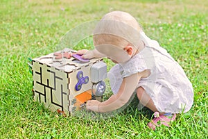 Cute little girl is playing with busiboard outdoors on green grass. Educational toy for toddlers. girl opened door to cube of
