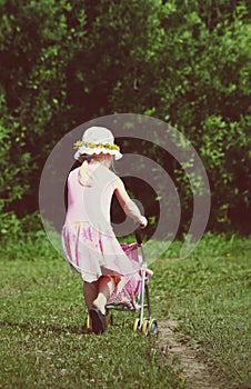 Cute little girl playing with her baby carriage on the meadow on sunny summer day.