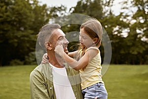 Cute little girl playing with her loving father while visiting park on a summer day, touching his cheeks and smiling