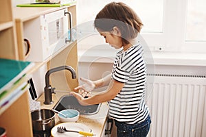 Cute little girl playing in her nursery room with toy child`s dishware, cooking food in toy kitchen