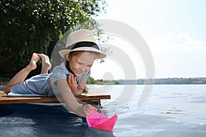 Cute little girl playing with paper boat on wooden pier near river