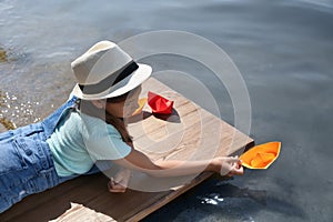 Cute little girl playing with paper boat on wooden pier near river