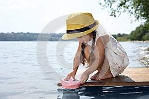 Cute little girl playing with paper boat on wooden pier near river
