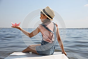 Cute little girl playing with paper boat on wooden pier near river